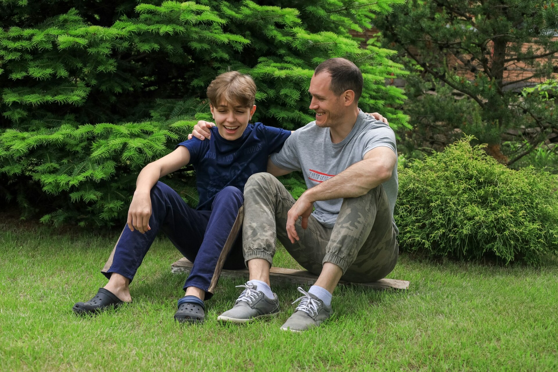 Dad is sitting on the lawn and hugging with his teen son at summer. The happy family is having fun